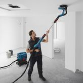 A person wearing safety equipment sands a ceiling with a drywall sander connected to a vacuum.