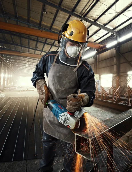 GWS 30-180 B A person wearing safety equipment grinds metal, producing bright sparks in a factory.