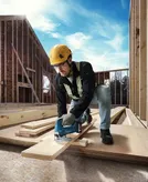 A person wearing safety equipment cuts a wooden plank with a cordless jigsaw at a construction site.