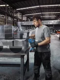 A person wearing safety equipment drills holes into metal air ducting in a factory.