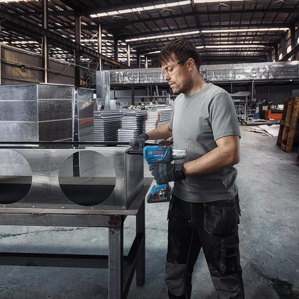 A person wearing safety equipment drills into a sheet metal duct in a factory.