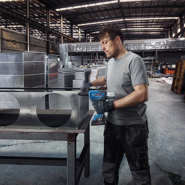 A person wearing safety equipment drills into a sheet metal duct in a factory.