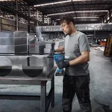 A person wearing safety equipment drills into a sheet metal duct in a factory.