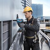A person wearing safety equipment uses a power drill to fasten panels on a building exterior.