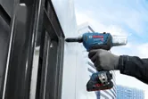 A person wearing safety equipment uses a cordless impact wrench on a steel frame.