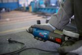 A person wearing safety equipment holds an angle grinder on a metal workbench.