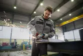 A person wearing safety equipment grinds a steel beam, producing flying sparks.