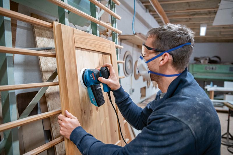 GEX 125-1 AE Person wearing safety equipment sanding a wooden cabinet door with a random orbit sander.