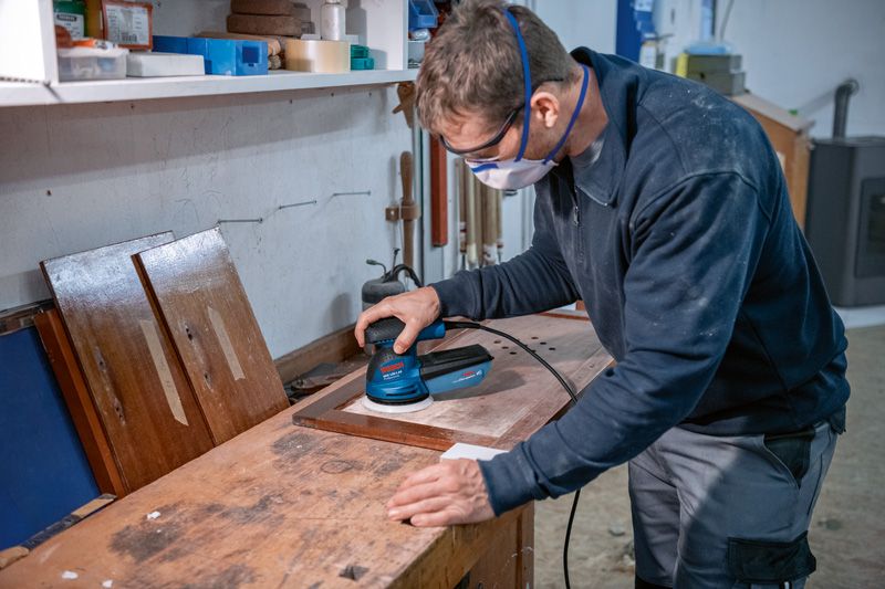 GEX 125-1 AE A person wearing safety equipment sands a wooden board with a random orbit sander.