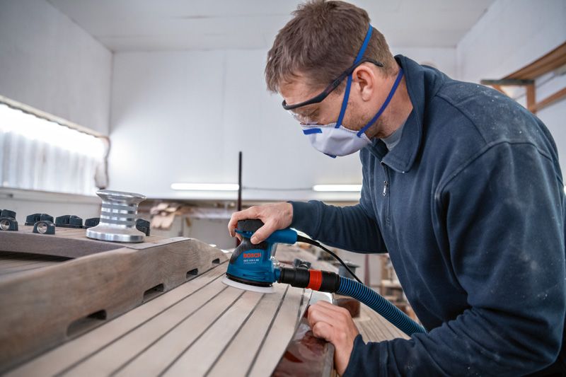 GEX 125-1 AE A person wearing safety equipment sands wooden planks with a random orbit sander.