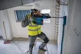 A person wearing safety equipment drills into a concrete wall using a cordless rotary hammer.