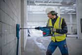 A person wearing safety equipment drills into a concrete wall using a cordless rotary hammer.