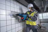 A person wearing safety equipment removes tiles from a wall with a cordless rotary hammer.
