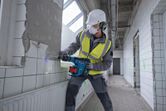 A person wearing safety equipment drills tiles on a wall in a tiled hallway.