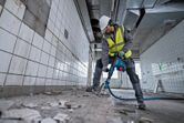 A person wearing safety equipment breaks up tile flooring with a cordless rotary hammer.