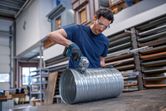 A person wearing safety equipment cuts a large metal duct with a cordless metal shear.