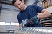 A person wearing safety equipment cuts a metal duct with a cordless metal shear.
