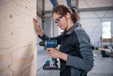 A person wearing safety equipment drills into a wooden board in a workshop.