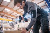 A person wearing safety equipment drills into wood with a cordless power tool.