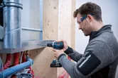 A person wearing safety equipment drills into a metal beam near exposed pipes.