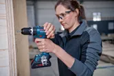 A person wearing safety equipment uses a cordless drill on a wooden panel.