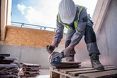 A worker wearing safety equipment uses a cordless angle grinder to cut tiles.
