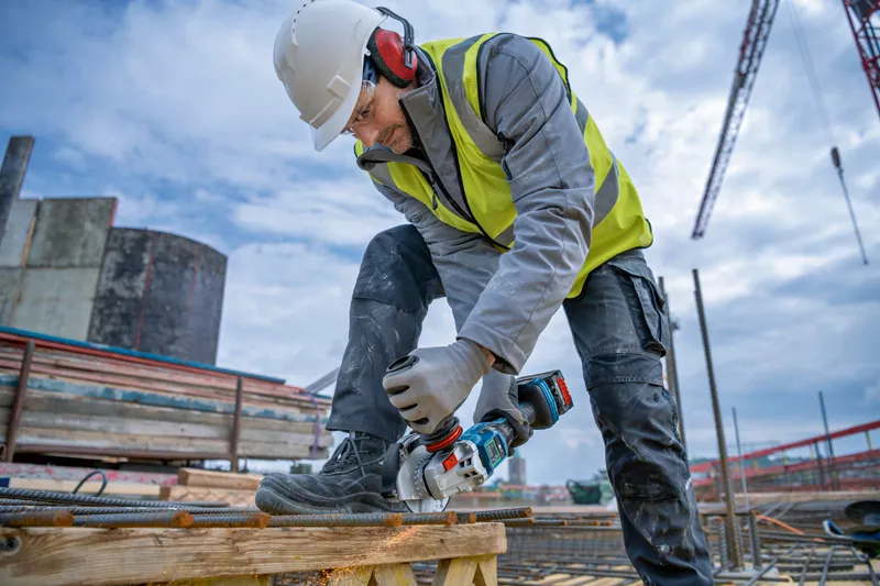 A person wearing safety equipment cuts rebar with a cordless angle grinder at a construction site.