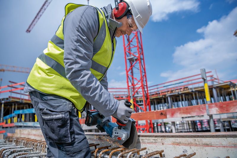 A person wearing safety equipment uses a cordless angle grinder to cut rebar at a construction site.