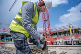 A person wearing safety equipment uses a cordless angle grinder to cut rebar at a construction site.