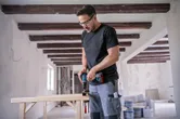 A person wearing safety equipment drills into wood at an indoor construction site.