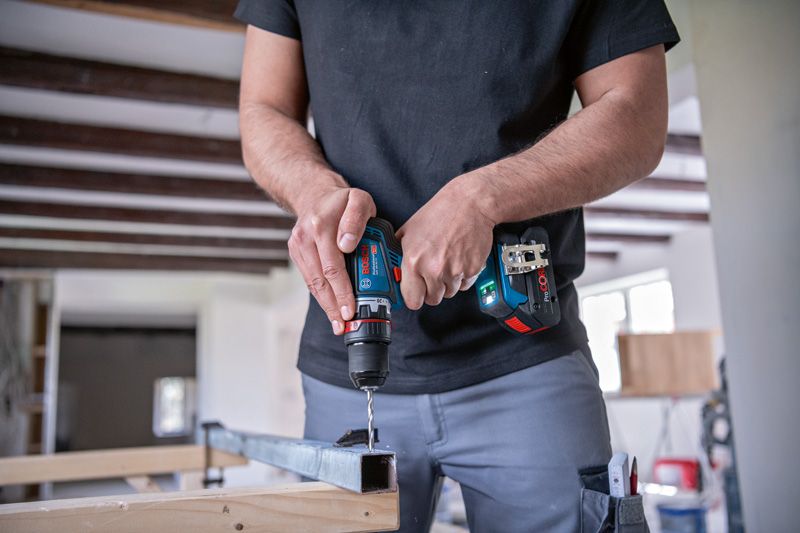 A person drills into a metal beam using a cordless driver inside a workshop.
