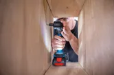 A person wearing safety equipment uses a cordless drill to drive a screw inside a wooden frame.