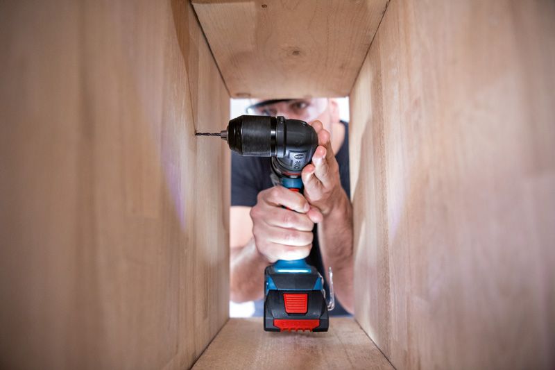 Person wearing safety equipment drills into a wooden panel inside a cabinet.