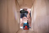 Person wearing safety equipment drills into a wooden panel inside a cabinet.