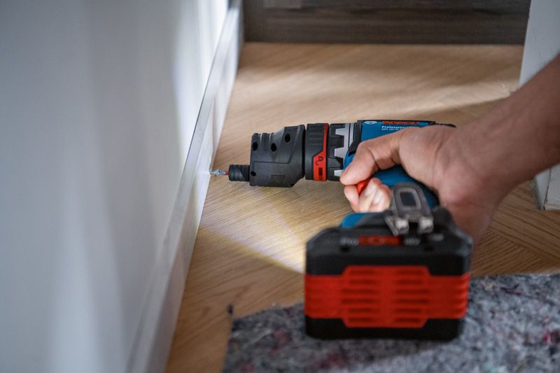 A person drills into a baseboard using a cordless drill on a wooden floor.