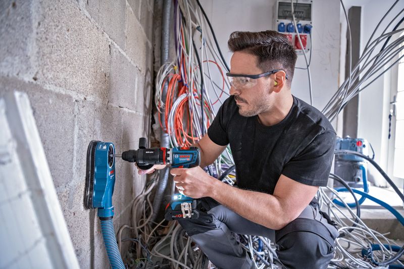 A person wearing safety equipment drills into a concrete wall with dust extraction.
