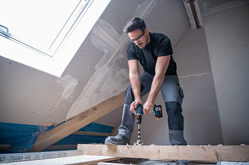 A person wearing safety equipment drills into a wooden beam in a partially finished attic.