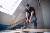 A person wearing safety equipment drills into a wooden beam in a partially finished attic.