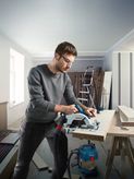 Person wearing safety equipment cuts wood with a cordless circular saw in a workshop.