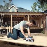 A person wearing safety equipment cuts wood with a cordless circular saw at a house construction site.