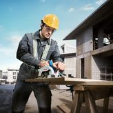 A person wearing safety equipment cuts wood with a cordless circular saw at a construction site.