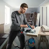 A person wearing safety equipment cuts wood with a cordless circular saw in a workshop.