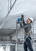 A person wearing safety equipment drills into a concrete ceiling while standing on scaffolding.