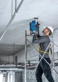 A person wearing safety equipment drills into a concrete ceiling while standing on scaffolding.