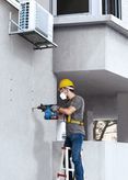 A person wearing safety equipment drills holes into a concrete wall beneath an air conditioner.