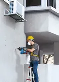 A person wearing safety equipment drills holes into a concrete wall beneath an air conditioner.