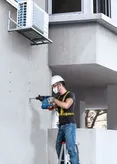 A person wearing safety equipment drills into a concrete wall beside an air conditioning unit.