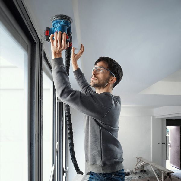 GEX 185-LI Person wearing safety equipment sands a ceiling near a window with a power sander.