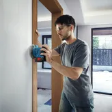 A person wearing safety equipment sands a wall with a cordless power sander.
