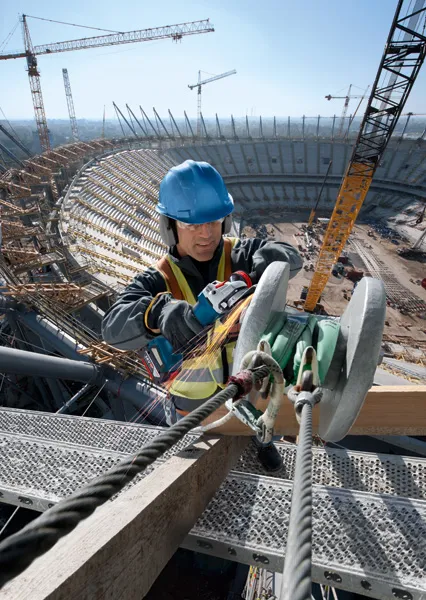 A person wearing safety equipment uses an angle grinder on a steel beam at a stadium construction site.